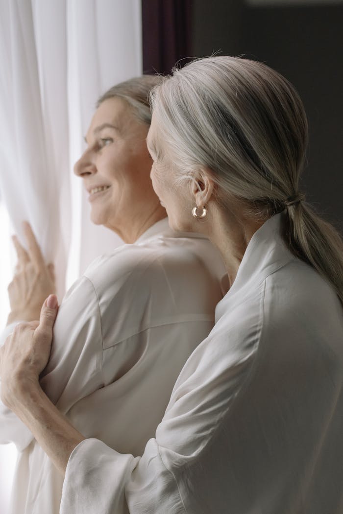 portfolio-04 Two senior women warmly embrace, enjoying a peaceful moment by a window.