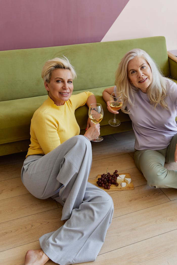portfolio-01 Two mature women enjoying white wine and cheese indoors, sitting casually on the floor.